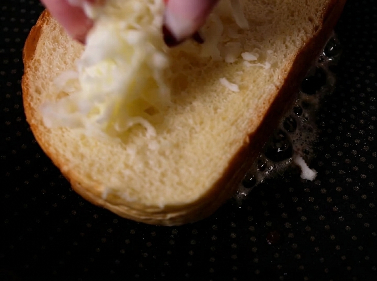 Dedos espolvoreando queso mozzarella rallado sobre una rebanada de pan en una sartén caliente.