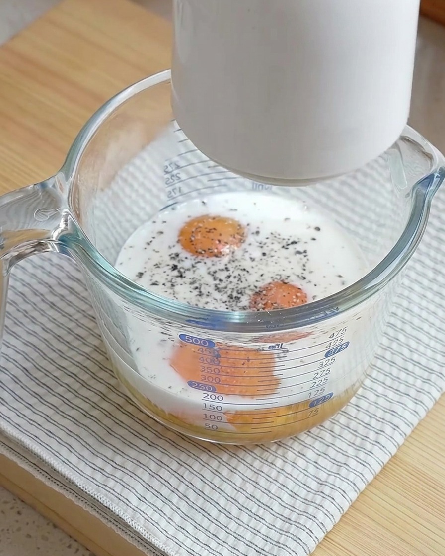 Un espumador blanco sostenido sobre una taza medidora de vidrio llena de huevos crudos, leche y pimienta negra recién molida.