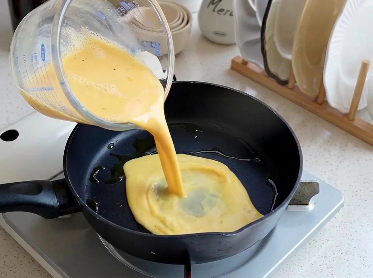 Mezcla amarilla de huevo batido siendo vertida desde una taza medidora de vidrio en una sartén negra ligeramente aceitada.