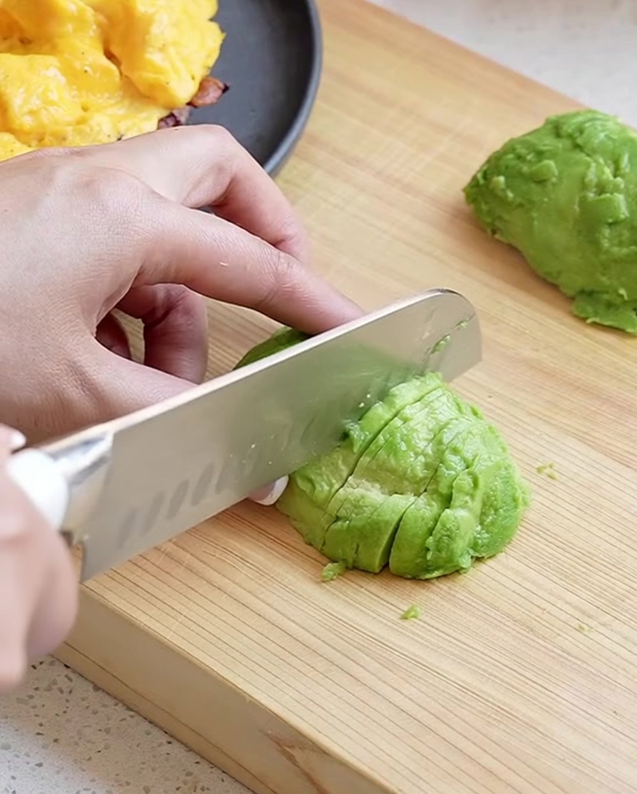 Una mano usando un cuchillo de chef para cortar la mitad de un aguacate pelado en tiras finas y ordenadas sobre una tabla de madera.