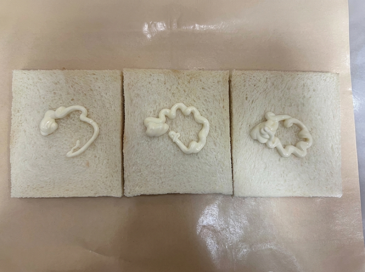 Tres rebanadas de pan blanco sin corteza colocadas una al lado de la otra sobre papel pergamino marrón, cada una con una línea en zig-zag de aderezo blanco.