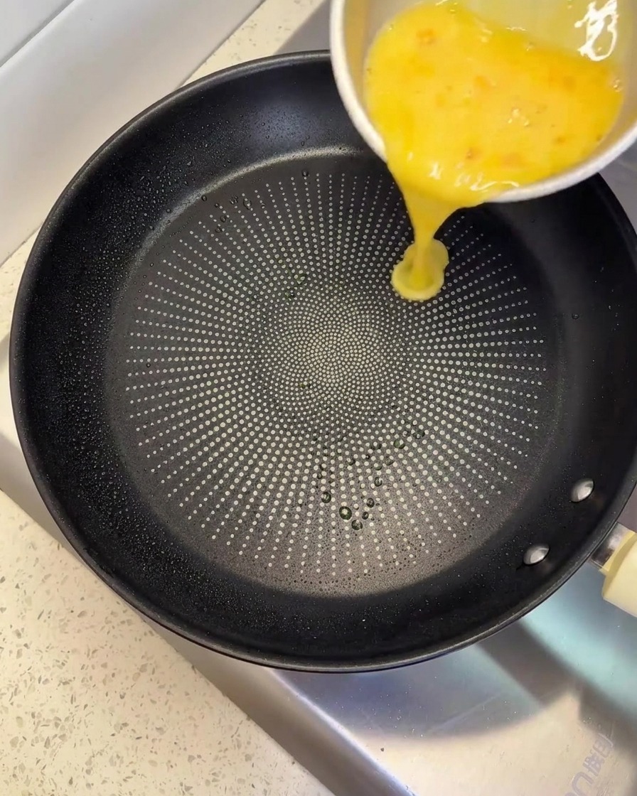 Vertiendo una mezcla de huevo batido amarillo desde un bol de metal hacia una sartén negra antiadherente.