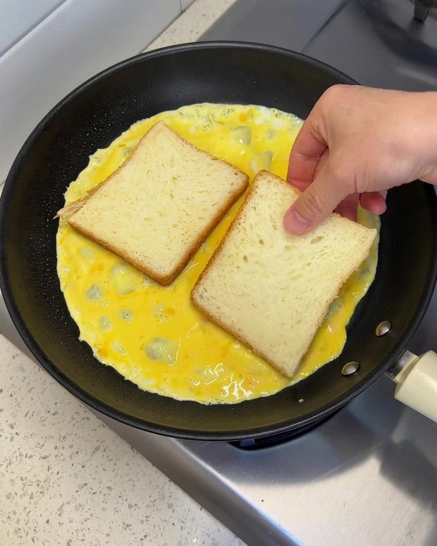 Una mano colocando dos rebanadas de pan blanco una al lado de la otra sobre una tortilla de huevo húmeda cocinándose en una sartén.