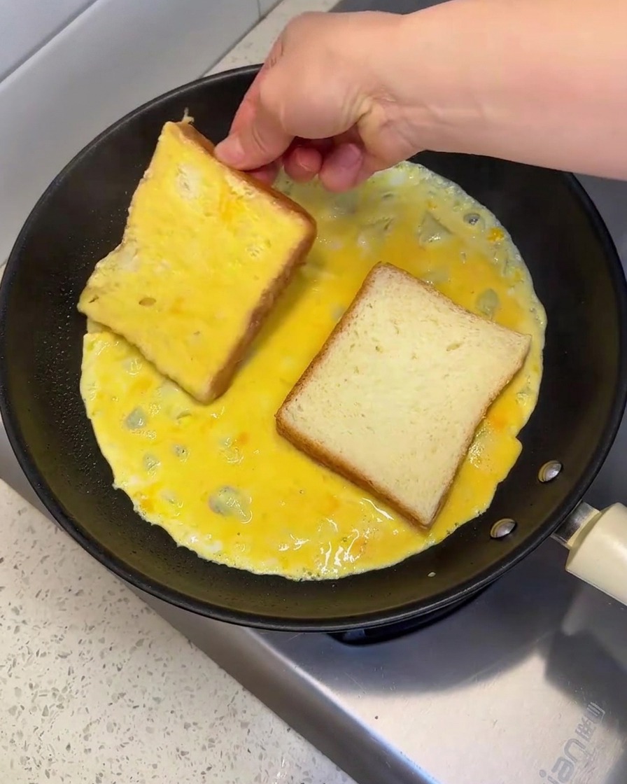Una mano colocando y volteando una rebanada de pan en una sartén caliente llena de huevo batido crudo para cubrir ambos lados.