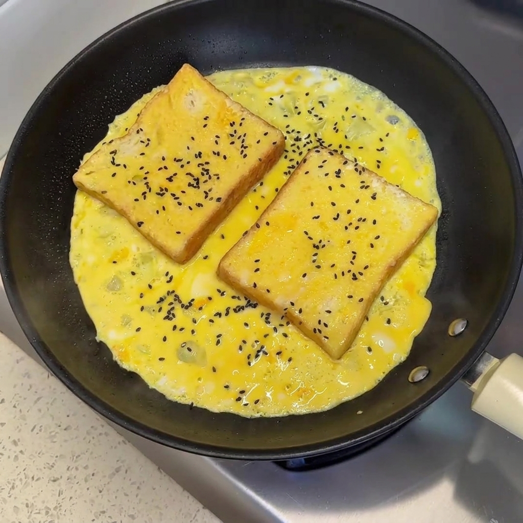 Semillas de sésamo negro espolvoreadas sobre dos rebanadas de pan ubicadas en una tortilla de huevo parcialmente cocida en una sartén.