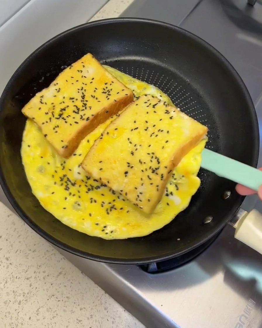 Una espátula de silicona levantando y volteando una gran tortilla de huevo con dos rebanadas de pan dentro en una sartén.