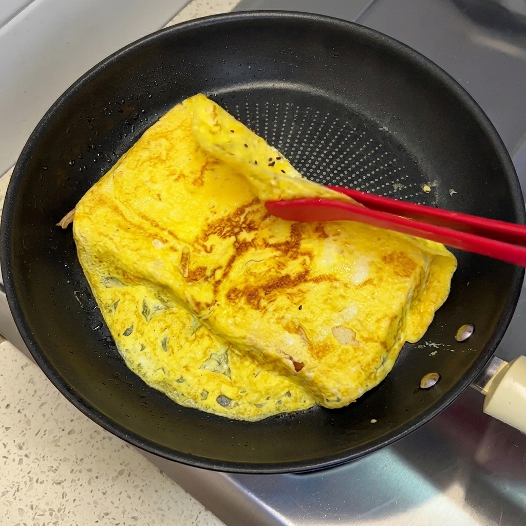 Pinzas de silicona roja doblando los bordes sobrantes de una tortilla de huevo cocida sobre dos rebanadas de pan en una sartén.