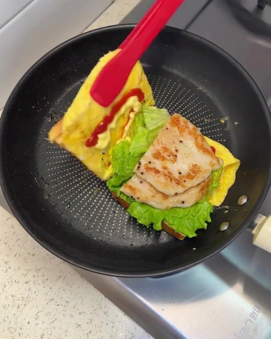Un par de pinzas rojas levantando y doblando un lado del pan para cerrar el sándwich en la sartén.