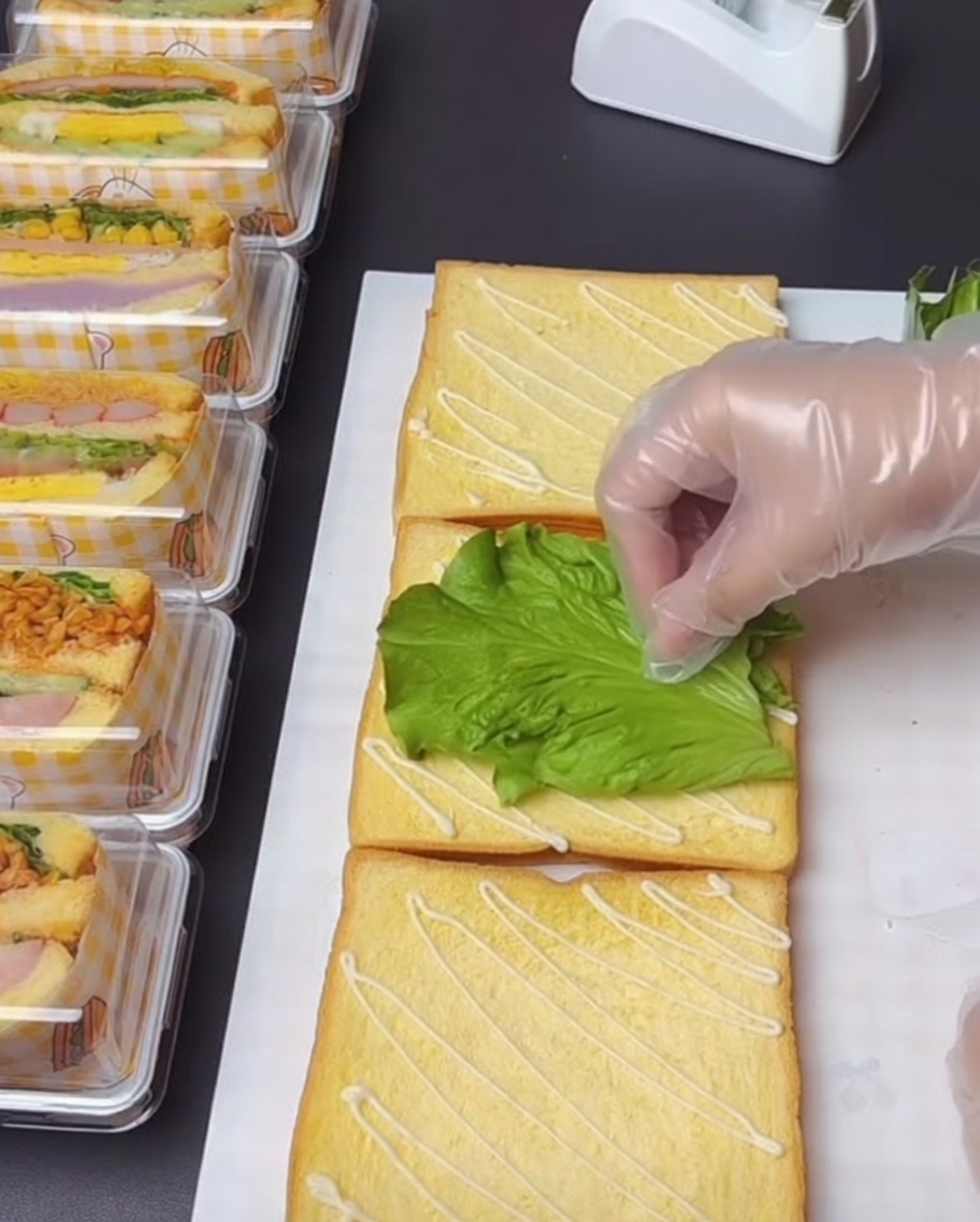 Una mano con guantes colocando cuidadosamente una hoja de lechuga verde fresca en una rebanada de tostada que ha sido rociada con aderezo de ensalada.
