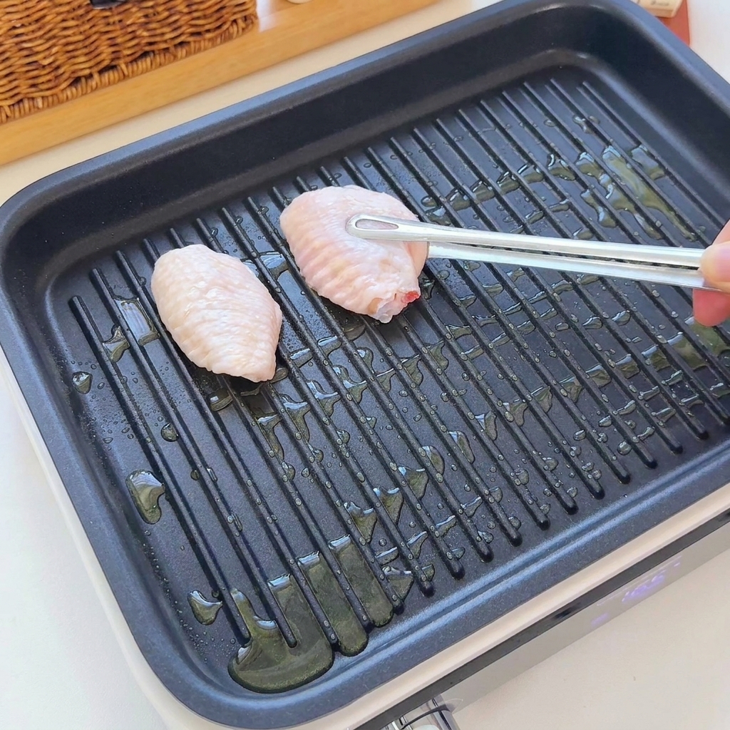 Alitas de pollo crudas siendo colocadas en una sartén de grill estriada con aceite usando pinzas.