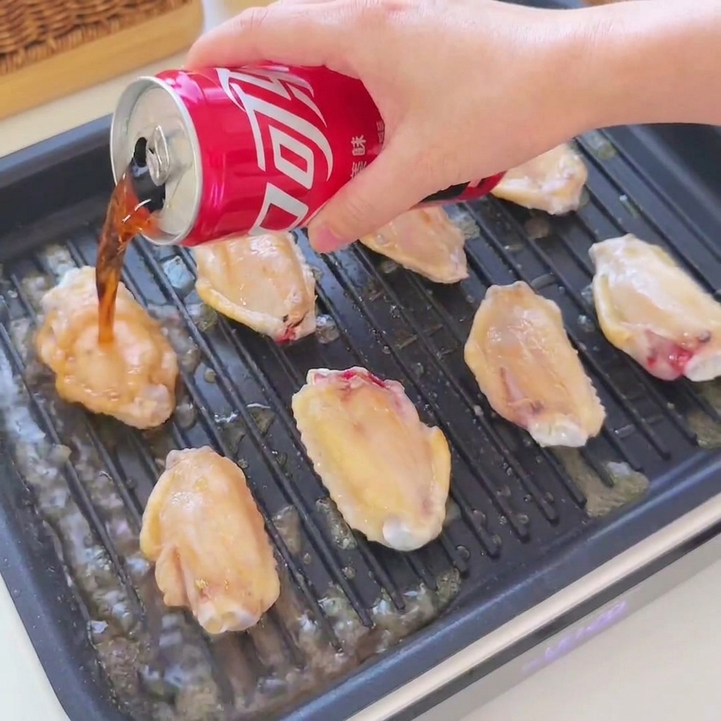 Cola y salsa de soja siendo vertidas desde una lata roja sobre alitas de pollo doradas chisporroteando en una sartén grill negra.