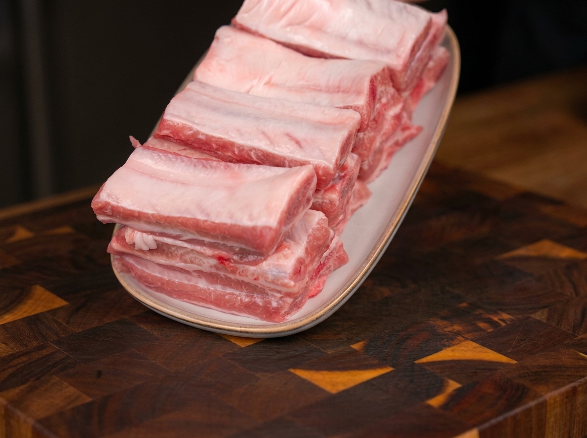 Costillas de cerdo crudas y frescas apiladas ordenadamente en un plato ovalado blanco, apoyadas sobre una tabla de cortar de madera.