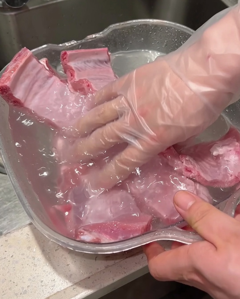 Manos con guantes lavando y fregando costillas de cerdo crudas dentro de un recipiente lleno de agua.