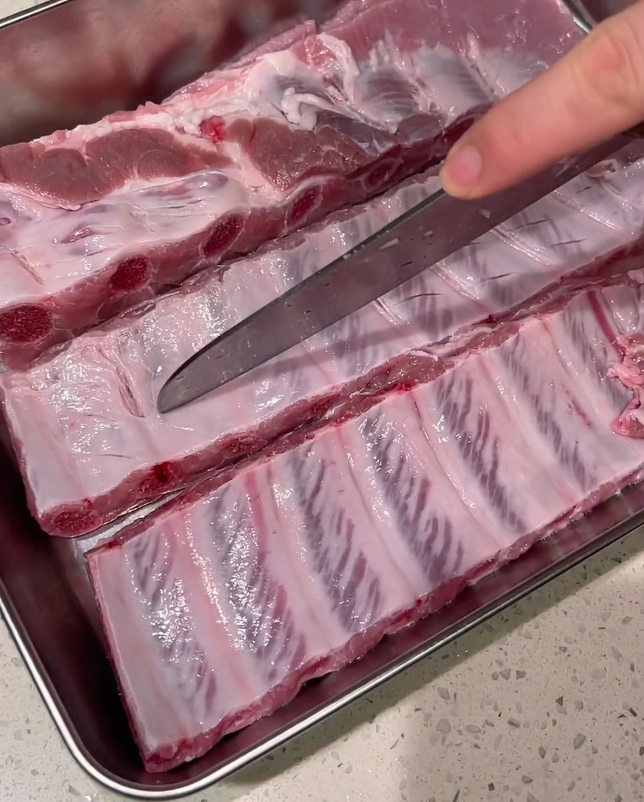 Un cuchillo afilado haciendo cortes poco profundos en la fascia de las costillas de cerdo crudas sobre una bandeja de metal.