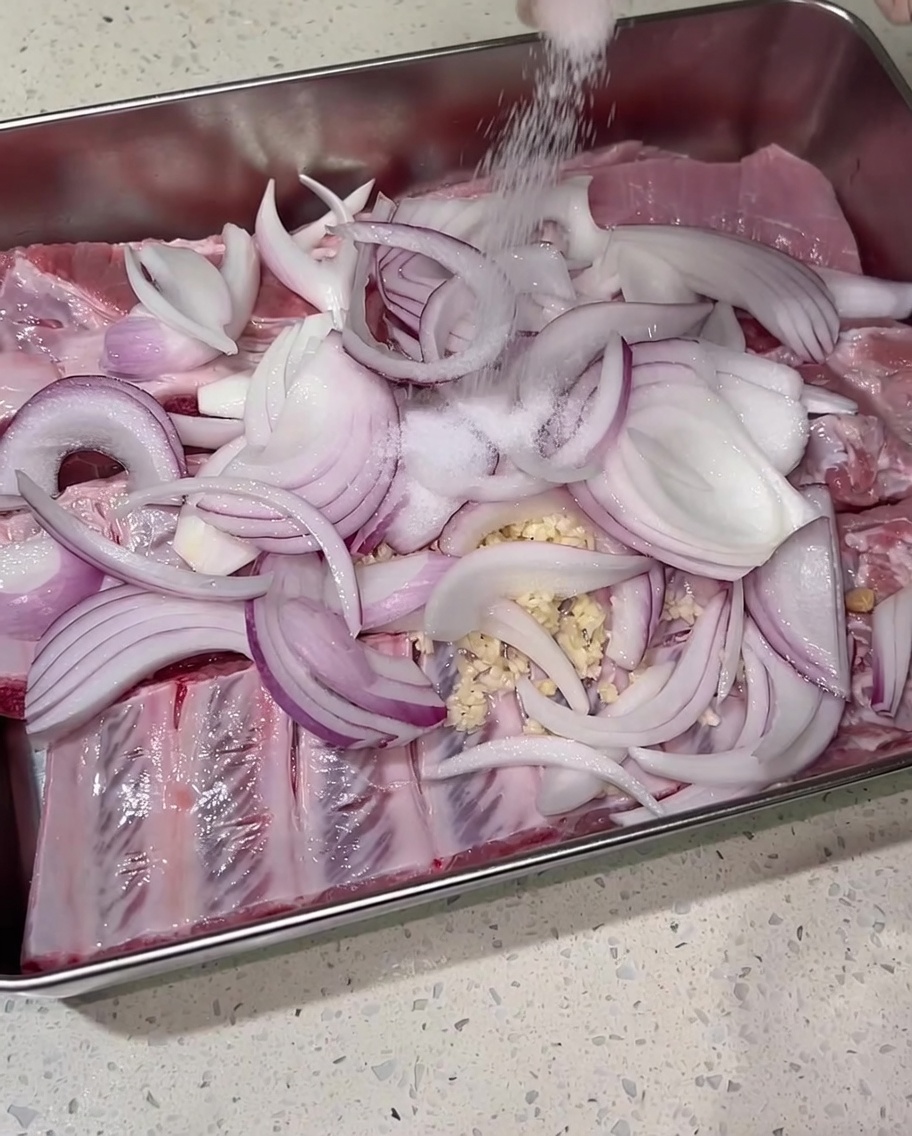 Sal siendo espolvoreada generosamente a mano sobre las costillas de cerdo sazonadas y las cebollas.