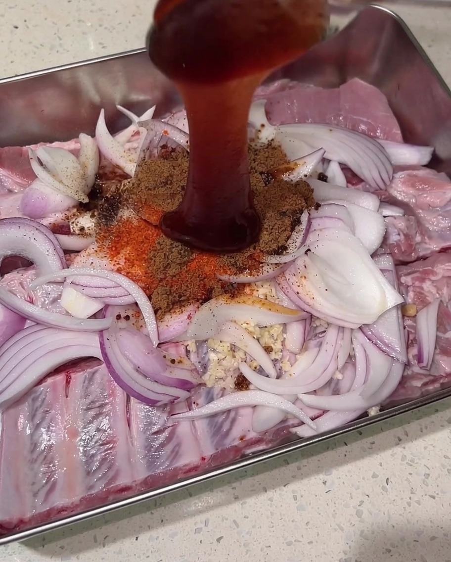 Salsa char siu espesa y de color rojo oscuro vertida desde arriba sobre el montón de especias secas, costillas de cerdo crudas y cebollas.
