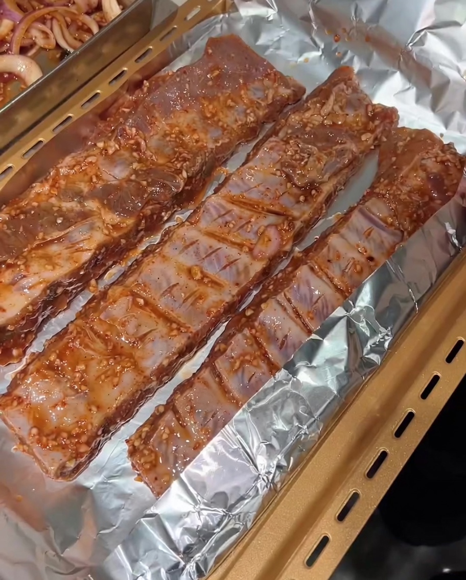 Tres tiras de costillas de cerdo marinadas colocadas sobre papel de aluminio en una bandeja para hornear dorada, listas para ser envueltas.