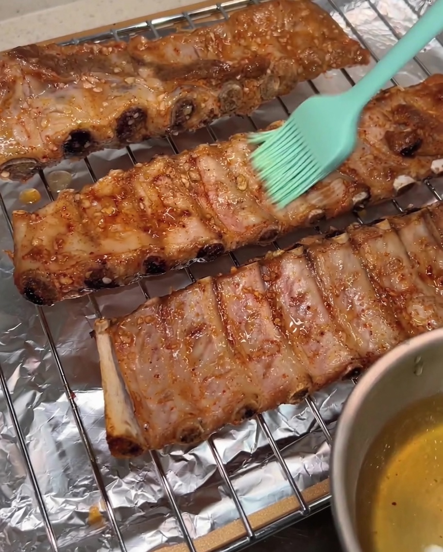 Un pincel de silicona verde aplicando un recubrimiento brillante de agua con miel sobre la superficie de las costillas de cerdo asadas en una rejilla.