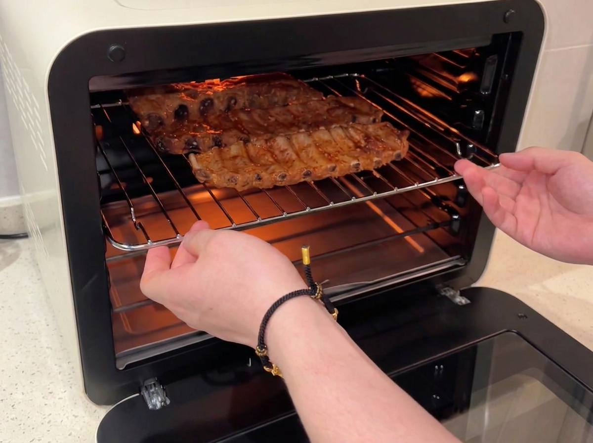 Manos deslizando una rejilla que sostiene costillas de cerdo glaseadas con miel de vuelta a un horno de convección encendido.