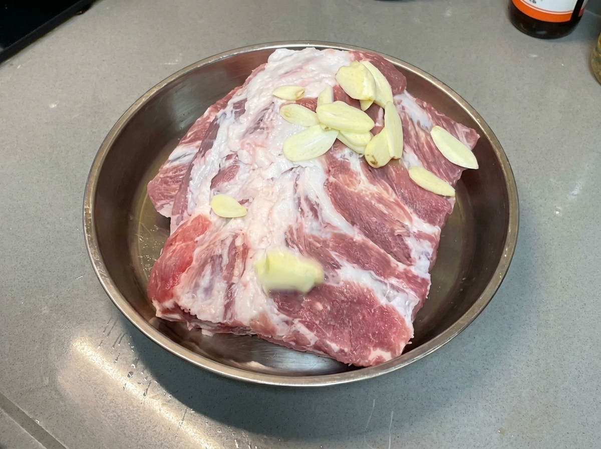 Rodajas de ajo esparcidas uniformemente sobre costillas de cerdo crudas en una bandeja de metal.
