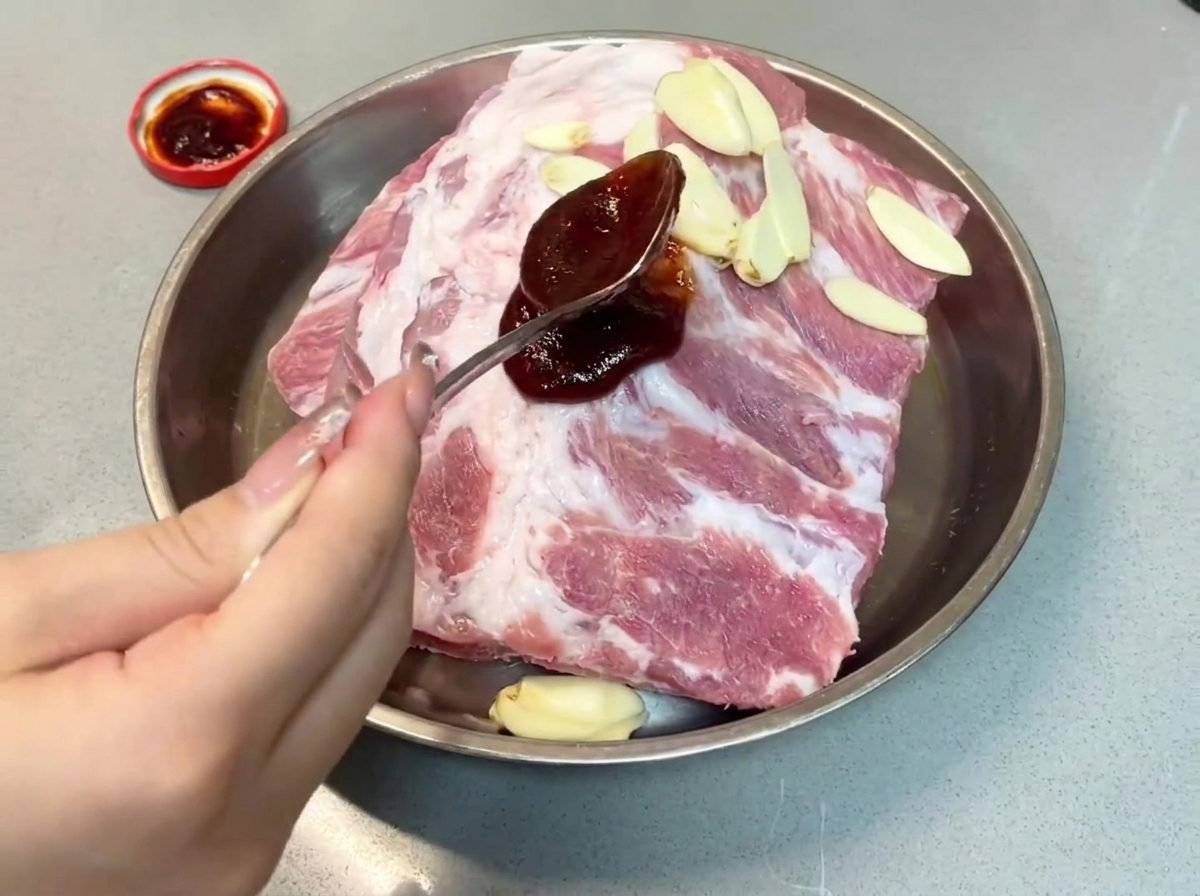 Una cucharada de salsa char siu oscura vertiéndose sobre costillas de cerdo en una bandeja de metal.