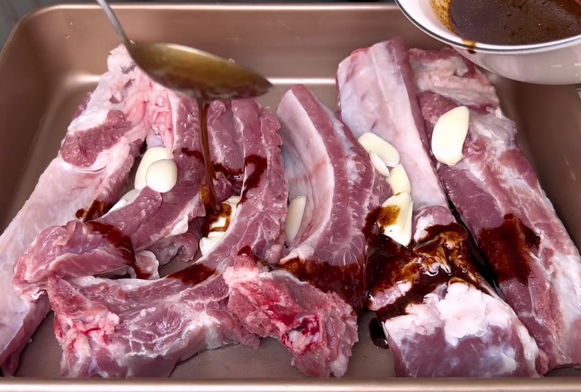 Vertiendo una marinada oscura a base de soja desde un tazón blanco sobre costillas de cerdo crudas y rodajas de ajo en una bandeja de horno.
