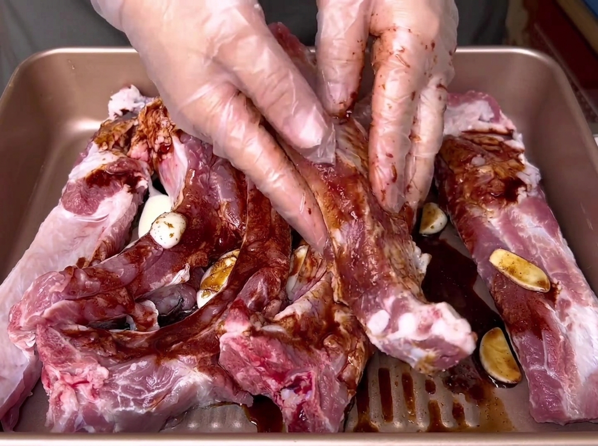 Manos enguantadas frotando a fondo una marinada líquida oscura en costillas de cerdo crudas dentro de una bandeja dorada.