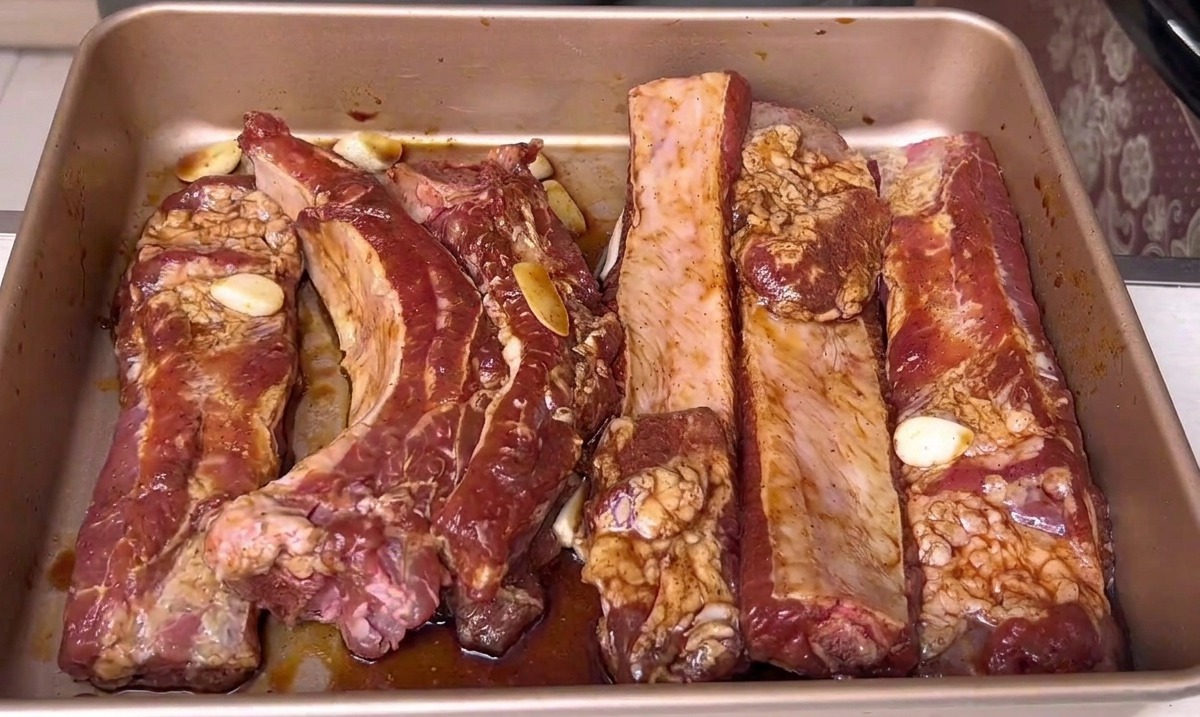 Costillas de cerdo marinadas descansando en una bandeja de horno dorada, listas para ser colocadas en el horno.