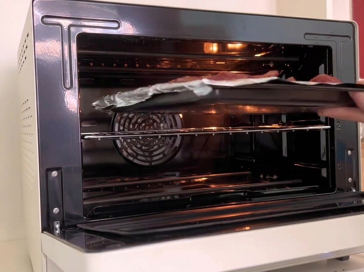 Una bandeja de horno que sostiene costillas envueltas en papel de aluminio siendo deslizada en la rejilla central de un horno blanco encendido.