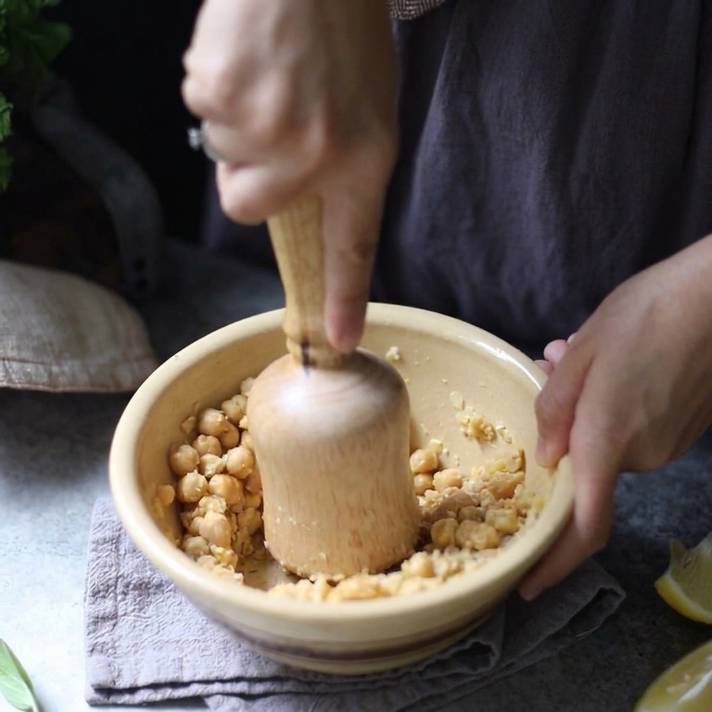 Una persona usando un mazo de madera para triturar manualmente garbanzos cocidos dentro de un cuenco de cerámica amarillo.