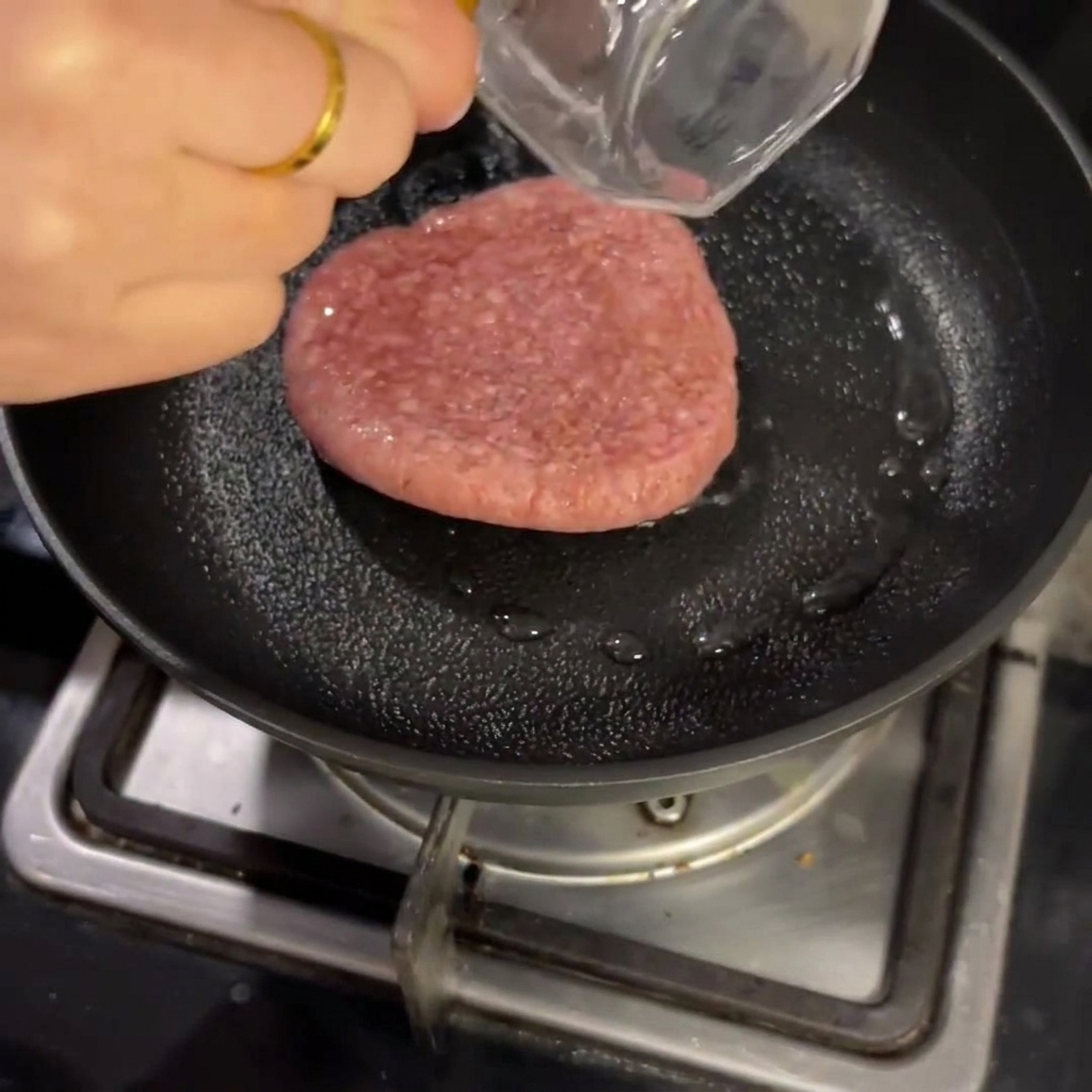 Medallón de carne cruda cocinándose en una sartén con un poco de líquido, cubierta con una tapa.
