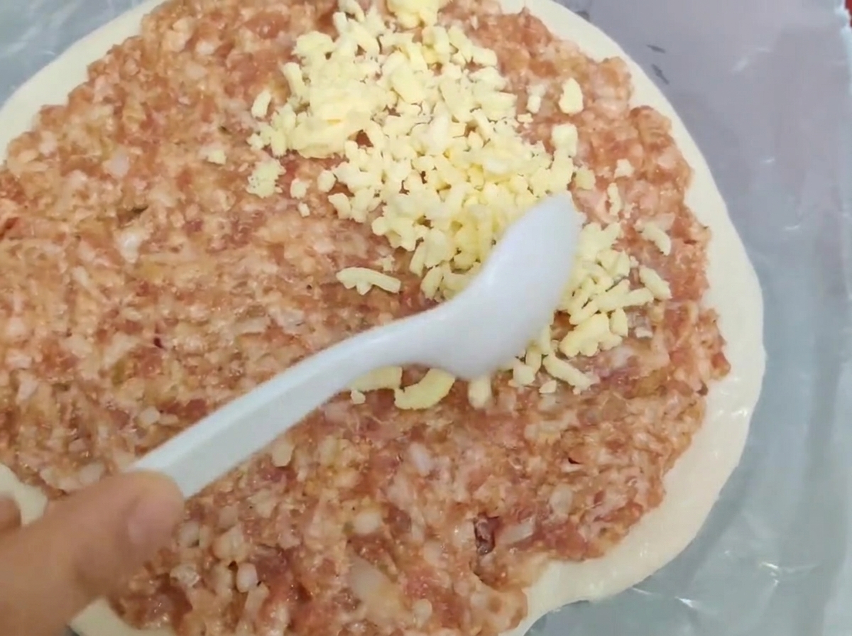 Una cuchara blanca extendiendo carne picada cruda y queso rallado sobre una base de masa redonda.