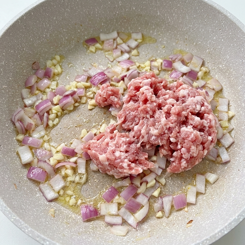 Carne de cerdo picada cruda en una sartén junto a cebollas cocinándose y ajo picado.