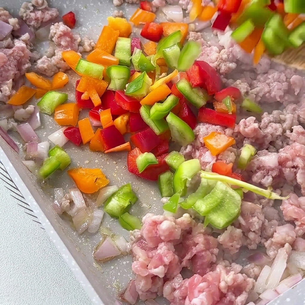 Pimientos mixtos en dados rojos, amarillos y verdes siendo agregados a la sartén con la mezcla de carne.