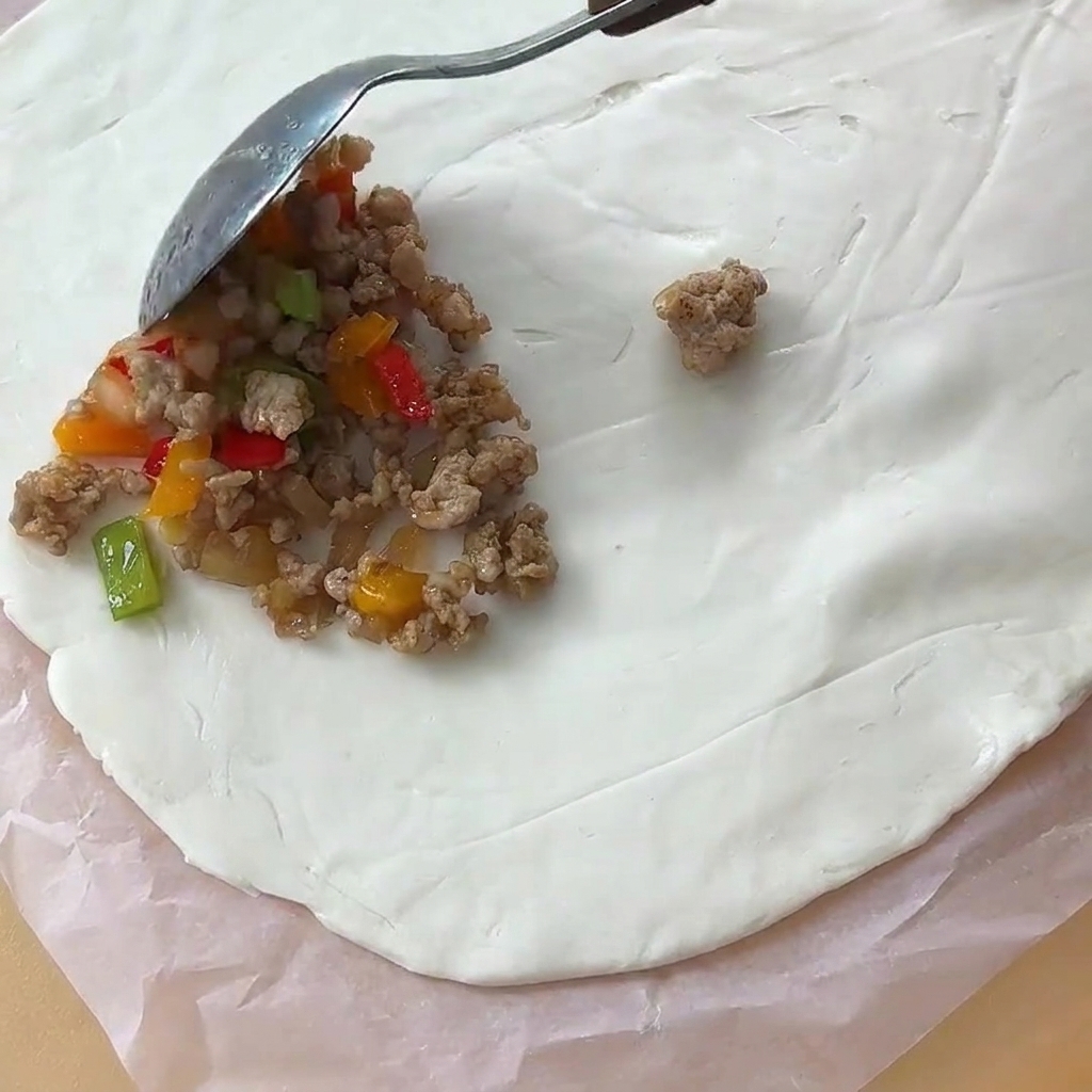 Una cucharada de relleno de carne cocida siendo colocada sobre una hoja circular de masa de pan plano en papel pergamino.