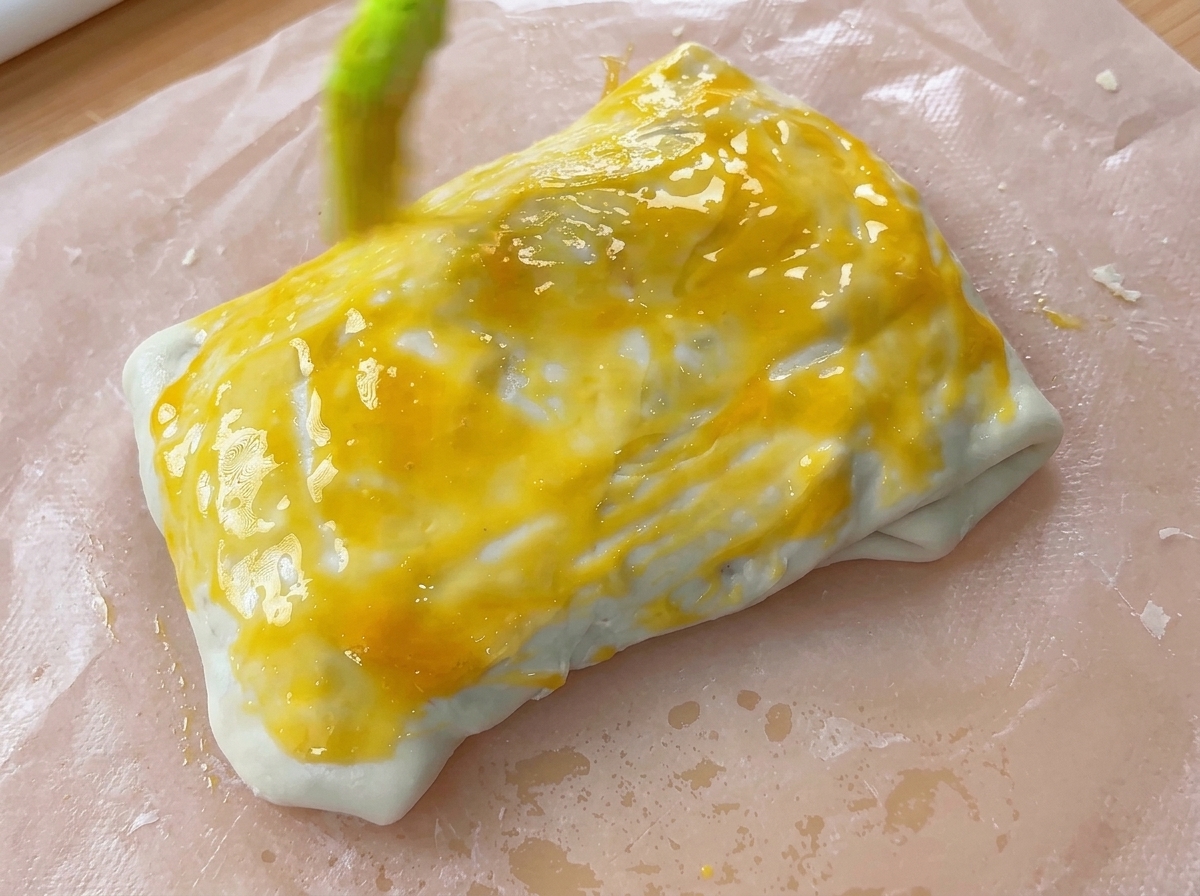 Un cepillo de repostería aplicando una capa de baño de huevo amarillo brillante sobre un paquete de masa doblada.