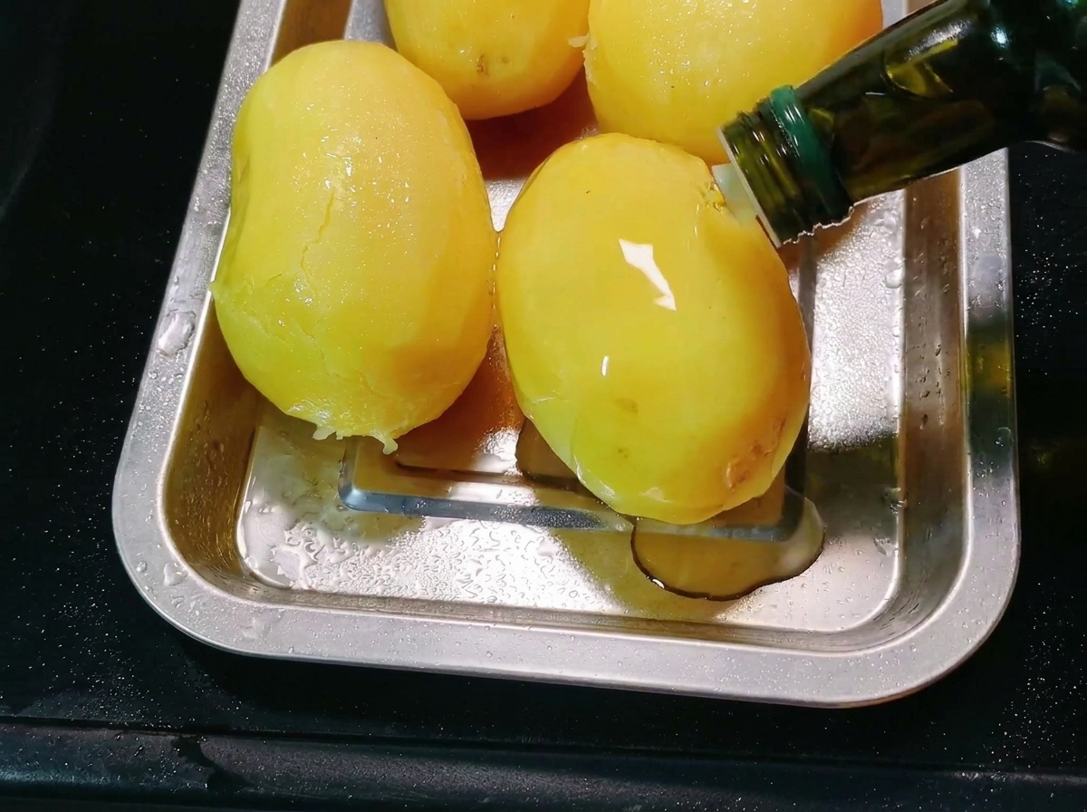 Aceite de cocina siendo vertido desde una botella sobre papas peladas en una bandeja de metal para hornear.