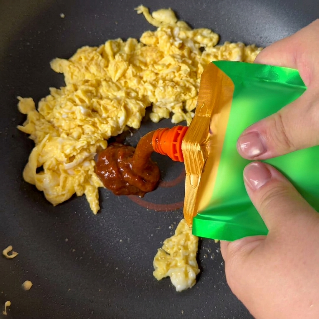 Pasta de soja amarilla exprimida desde una bolsa verde sobre los huevos revueltos en una sartén.