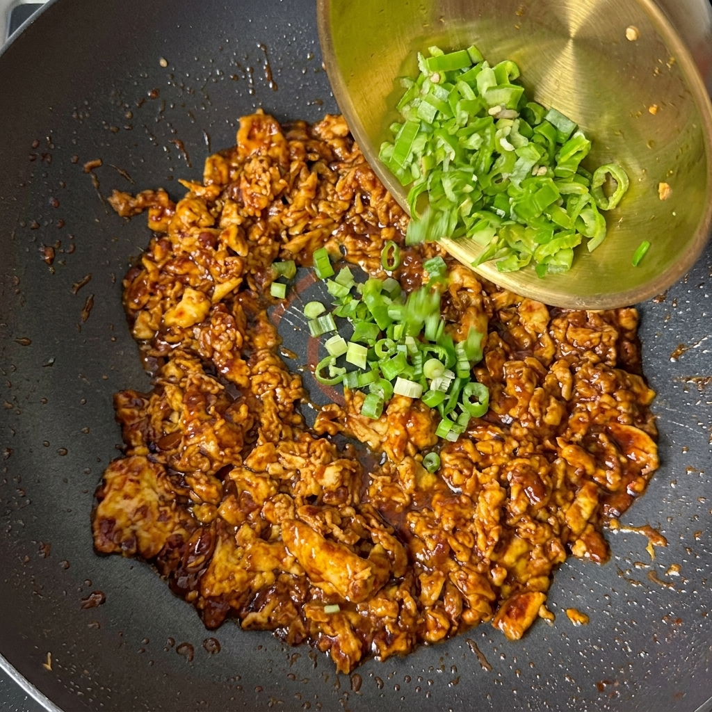 Cebollinos picados y trozos de pimiento verde vertidos desde un bol dorado sobre la mezcla de huevo cocido.