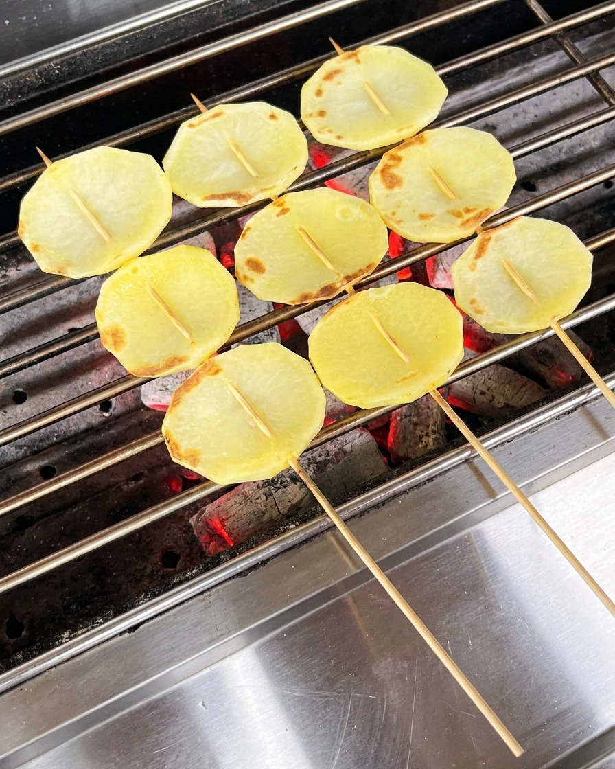Rodajas de patata parcialmente cocinadas en la parrilla siendo cubiertas con aceite usando un pincel.