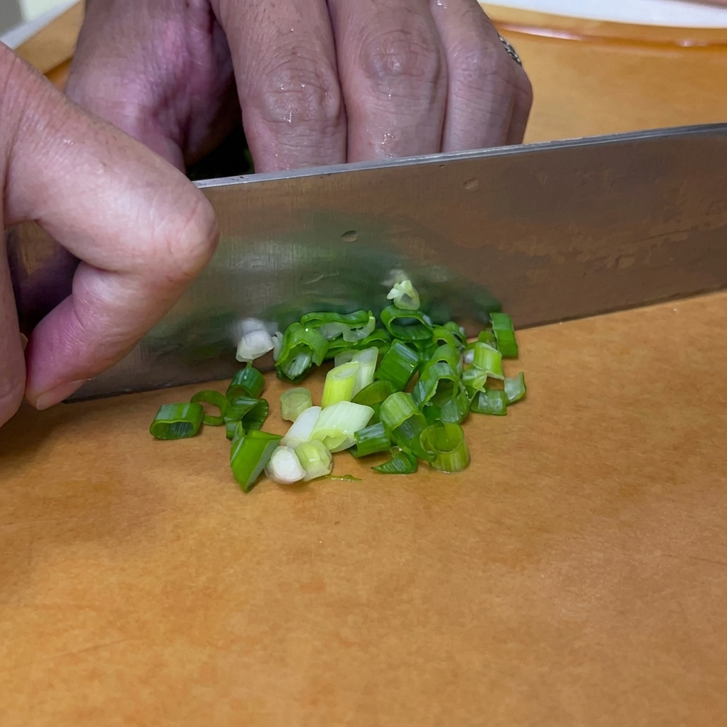 Cebollinos siendo picados en trozos pequeños y redondos con un cuchillo sobre una tabla de cortar.