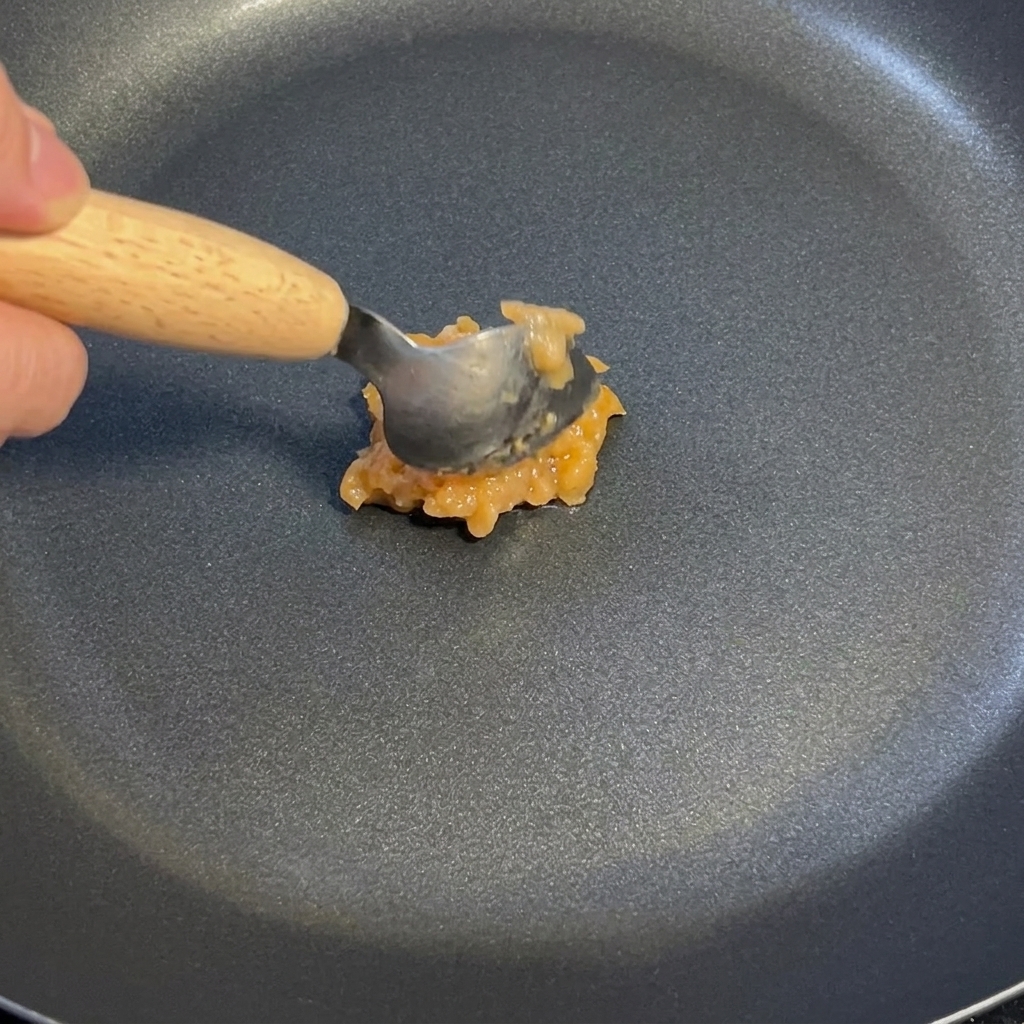 Una cucharada de salsa de ajo y jengibre siendo revuelta en una sartén antiadherente oscura a fuego lento.