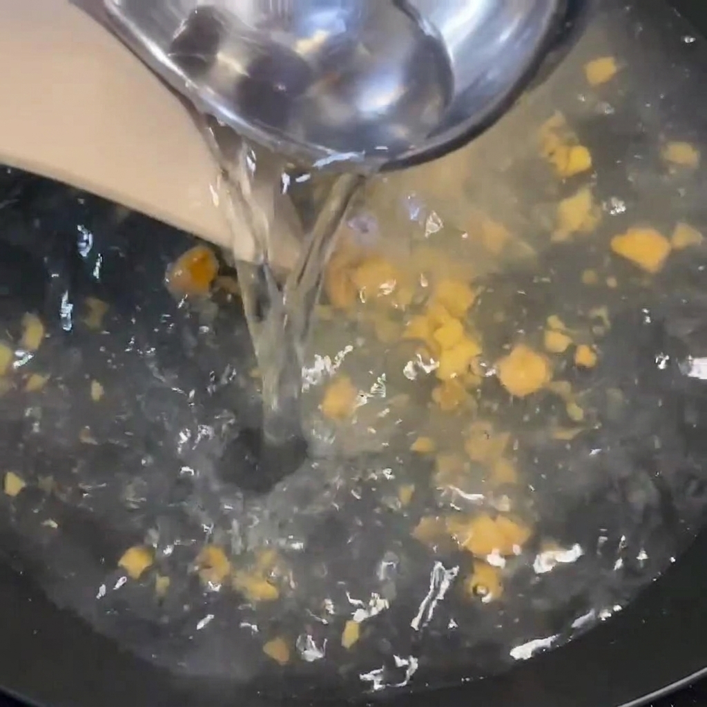 Agua caliente siendo vertida desde un cucharón en una sartén hirviendo que contiene condimento y trocitos de jengibre.