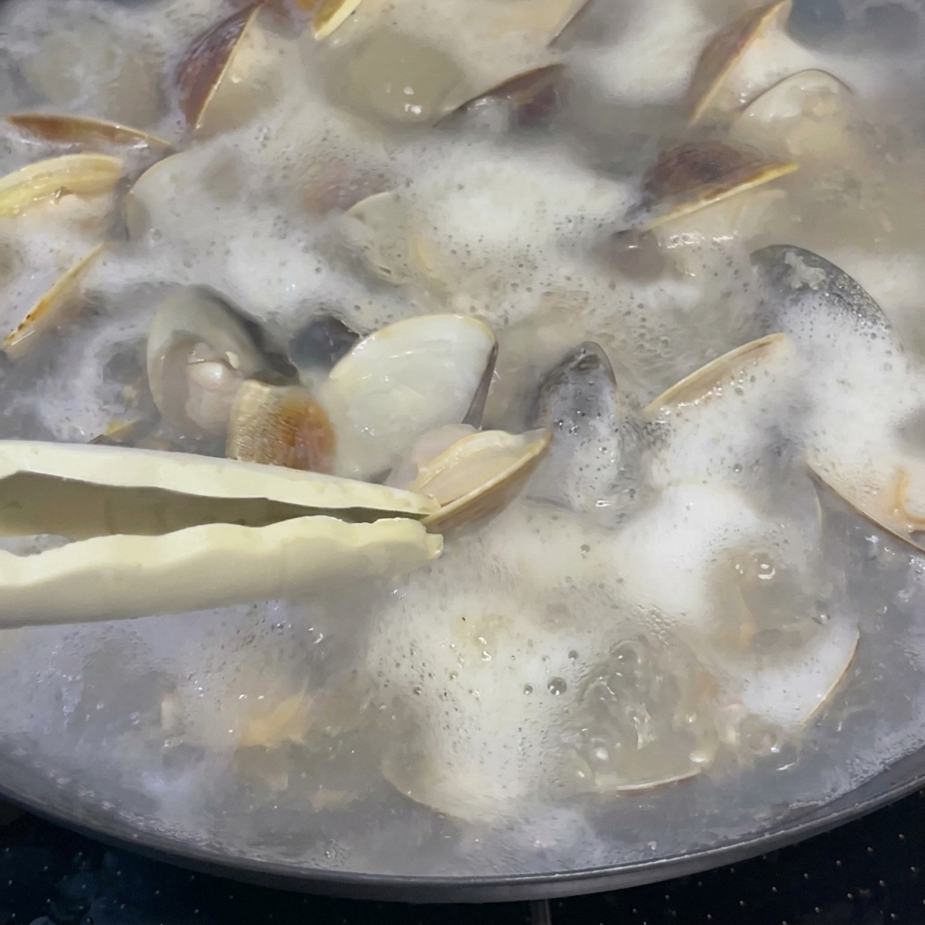 Una cucharada de vino de arroz siendo vertida en una olla de almejas y caldo burbujeantes.