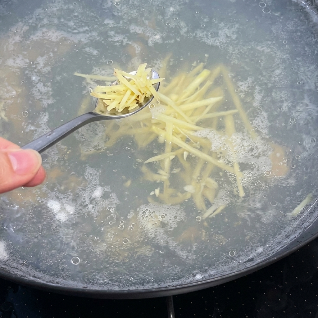 Una cucharada de jengibre julianizado siendo añadida a una olla hirviendo de caldo de sopa claro.