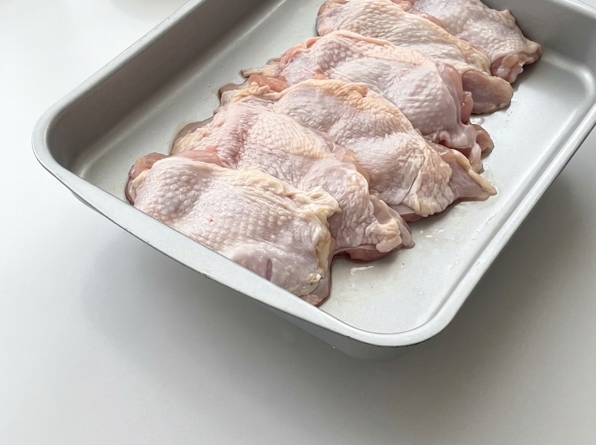 Muslos de pollo crudos dispuestos en una bandeja de metal con rodajas de jengibre fresco colocadas encima.