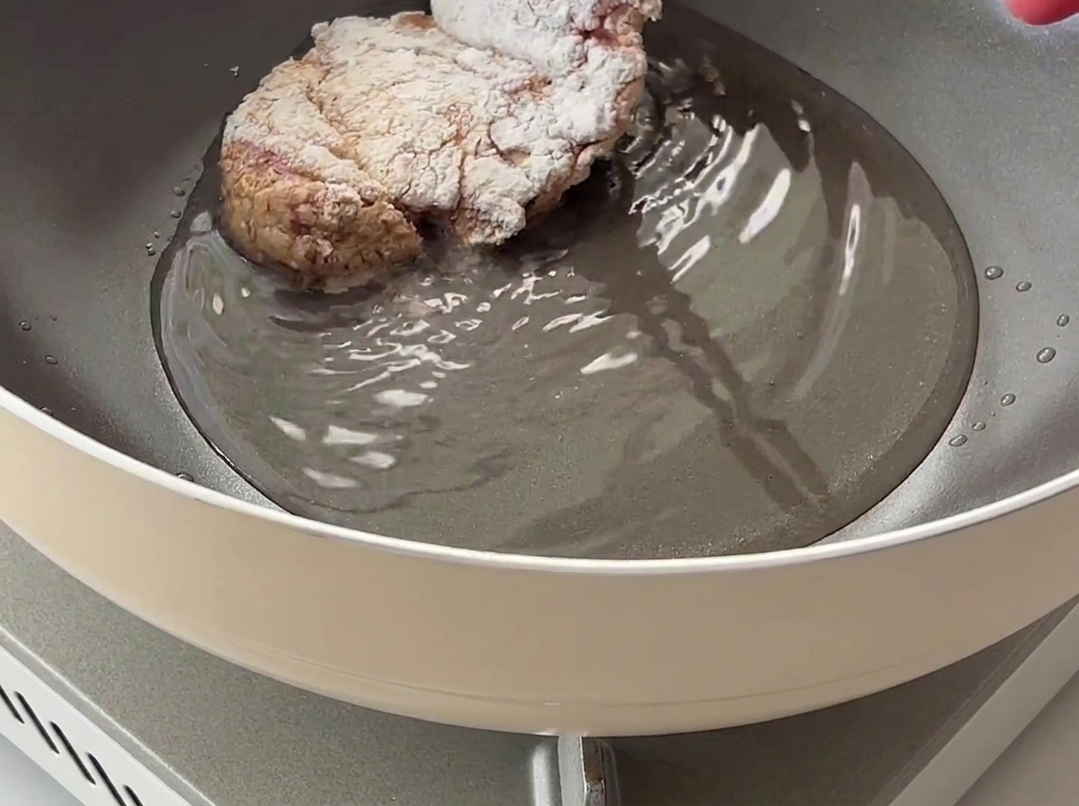 Un muslo de pollo cubierto de almidón de maíz siendo colocado en una sartén con aceite caliente.