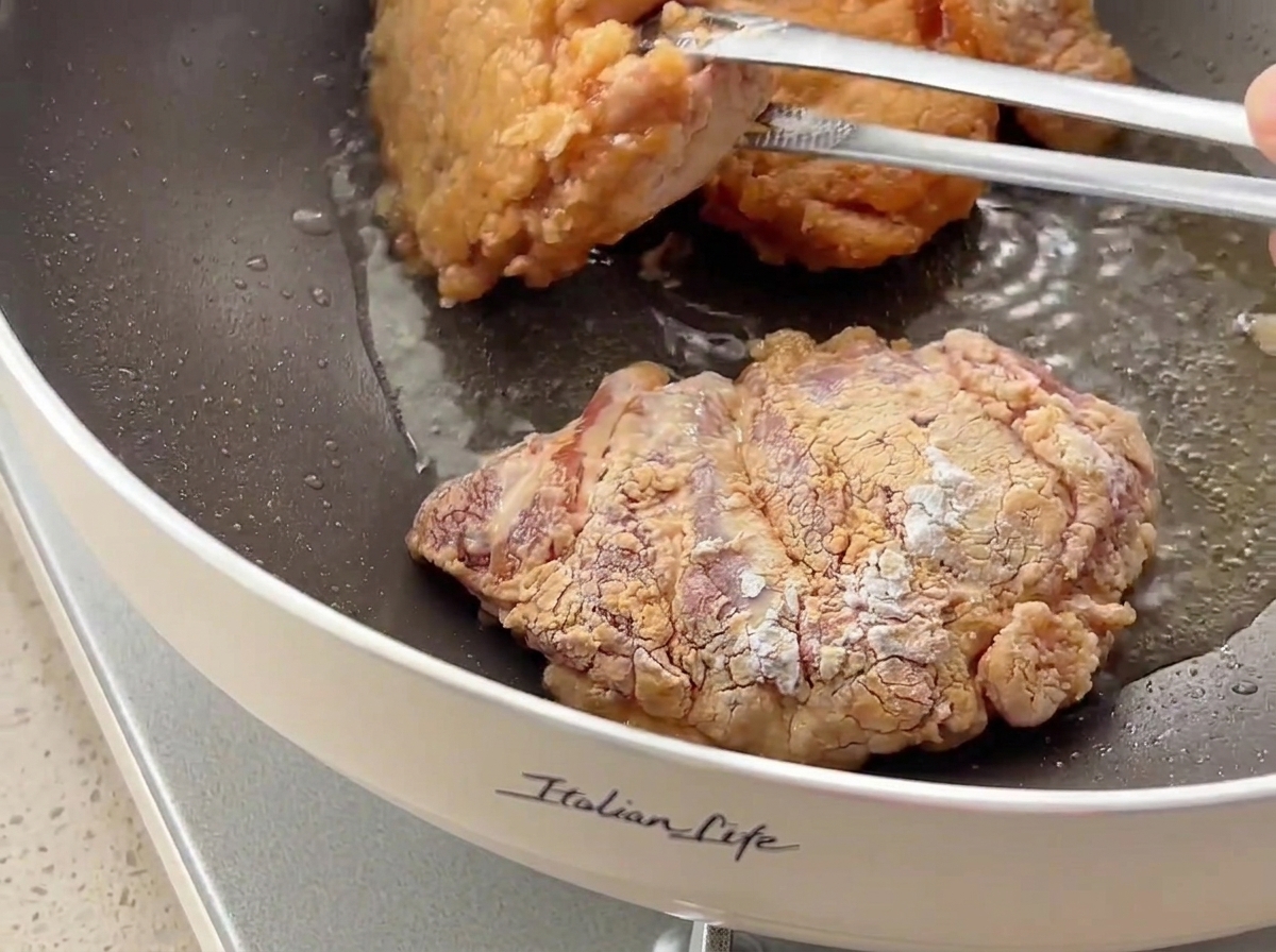 Piezas de pollo siendo volteadas con pinzas en una sartén, mostrando una costra dorada y crujiente.