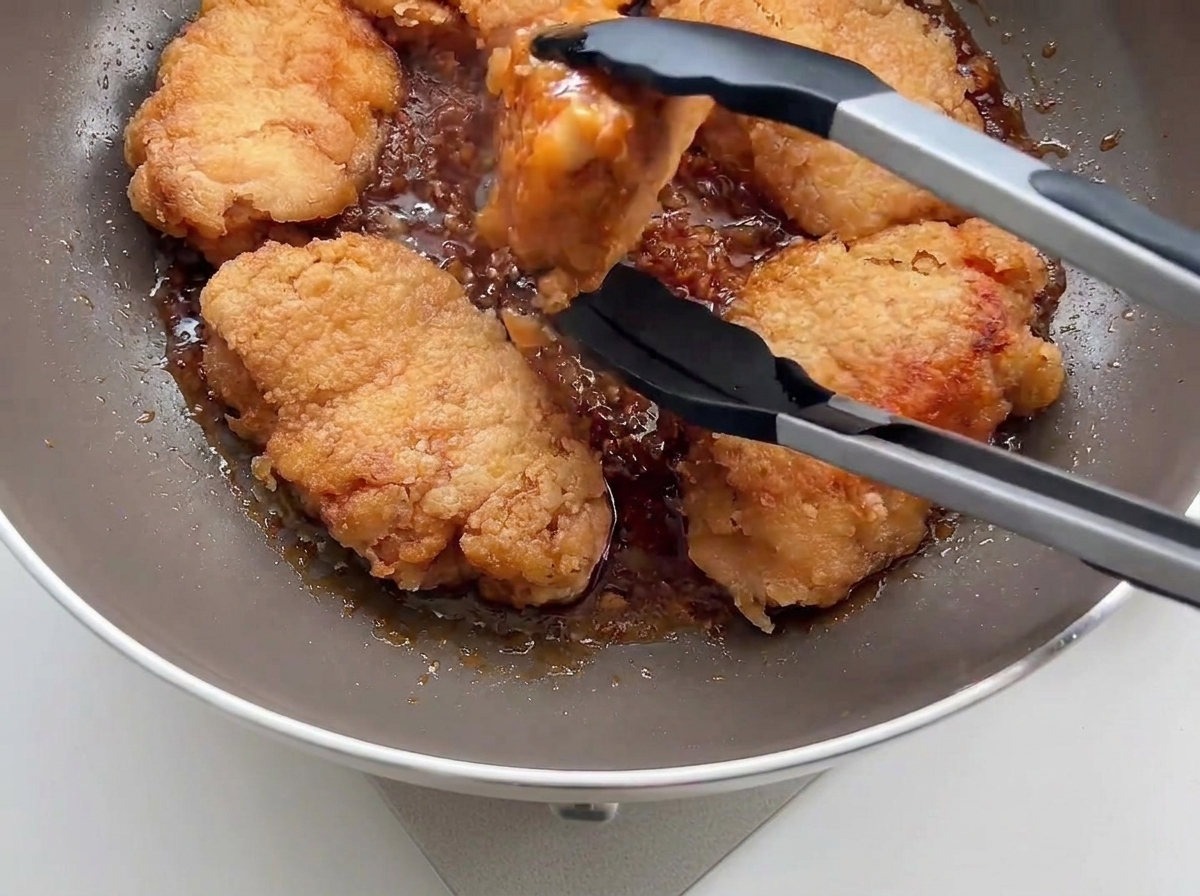 Pollo frito crujiente siendo colocado de nuevo en una sartén con salsa de ajo usando pinzas de cocina negras.
