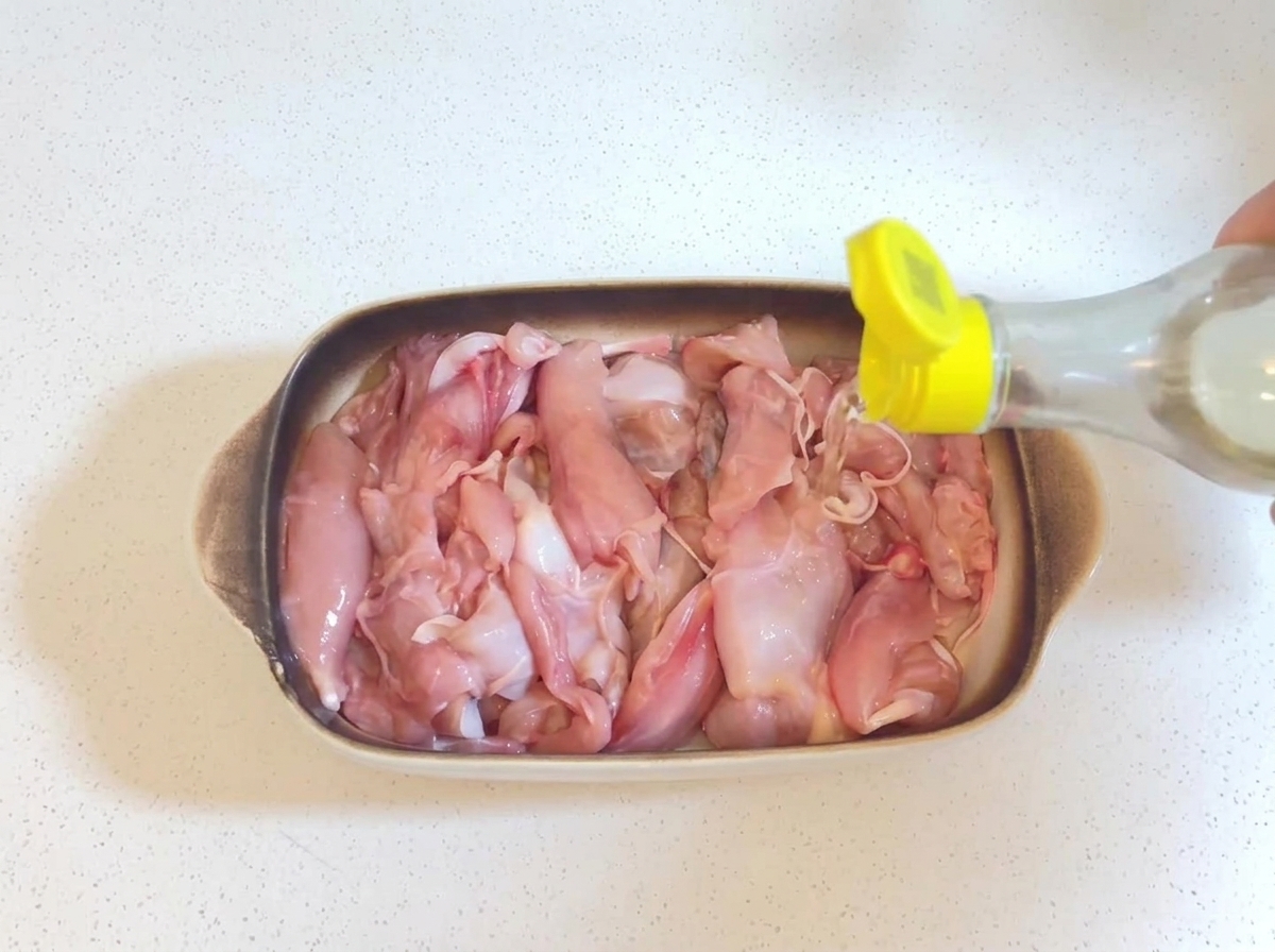 Vino de cocina transparente siendo vertido desde una botella pequeña con tapa amarilla sobre trozos de pollo crudo en una fuente para hornear.